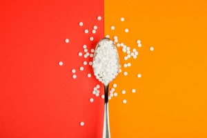 A bright red background on the left half of the picture with a bright orange background on the right side of the photo. There is a spoon full of artificial sweetener tablets in and around the spoon.