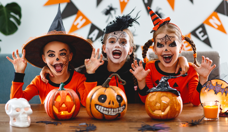 Three kids dressed up as witches with pumpkins for Halloween as they smile at the camera.