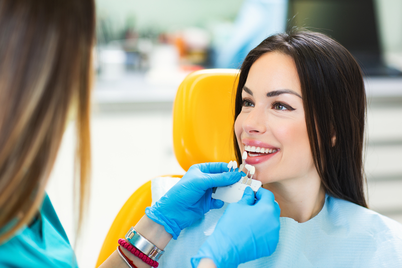 Young woman smiling in dental chair while the dentist holds different colored crowns to her teeth