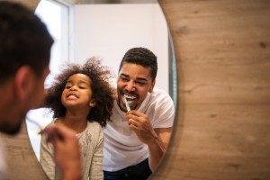 A young dad brushing his teeth in the mirror with his young daughter.